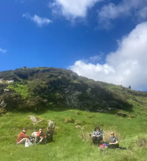 Five people in a sunny, sheltered picnic spot at the foot of a hill.